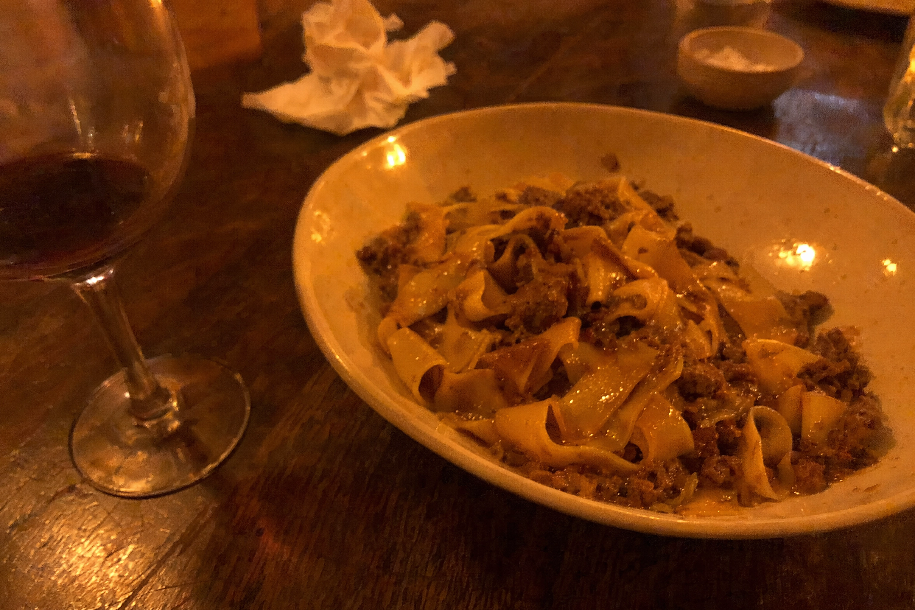 Bowl of pappardelle lamb ragu under a warm ceiling bulb, the orange cast that makes phone food photos read brown
