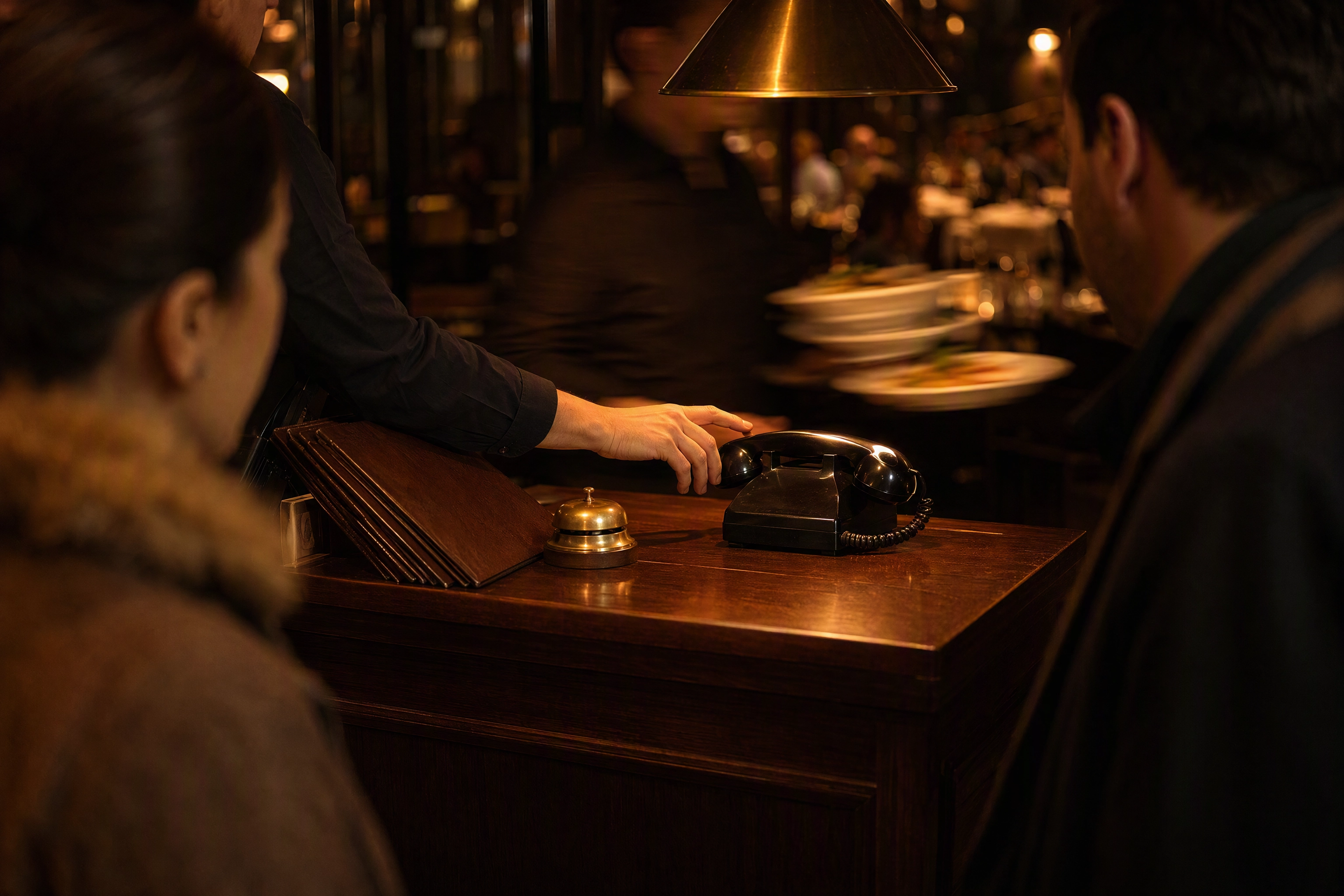 Host reaching for a ringing phone at a busy restaurant host stand, the front-of-house job AI voice answering tools replace