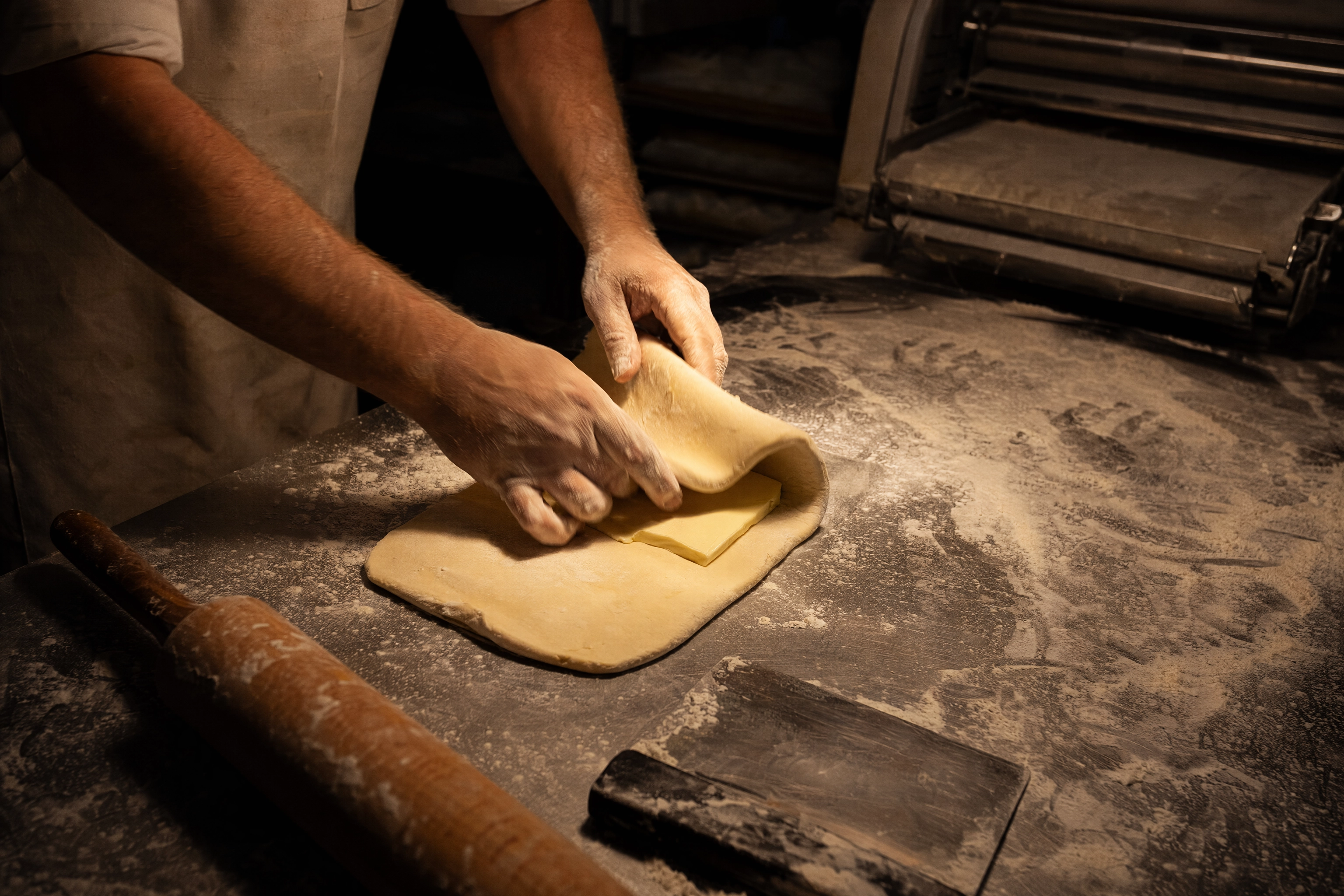 Baker folding a butter block into dough on a flour-dusted stainless bench during the 4am baking shift