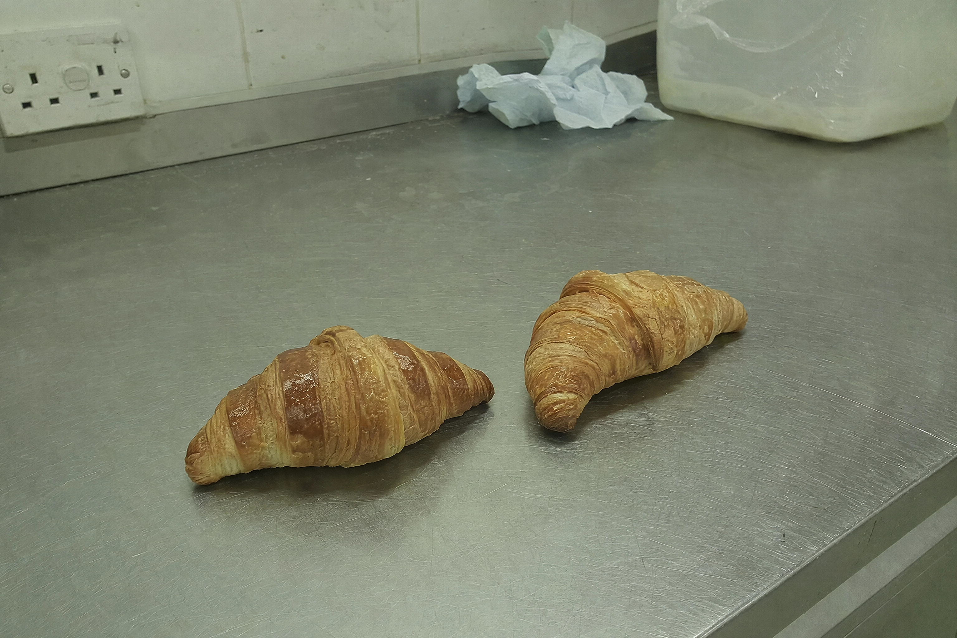 Flat unflattering phone snap of croissants on a stainless bench under fluorescent light before AI styling