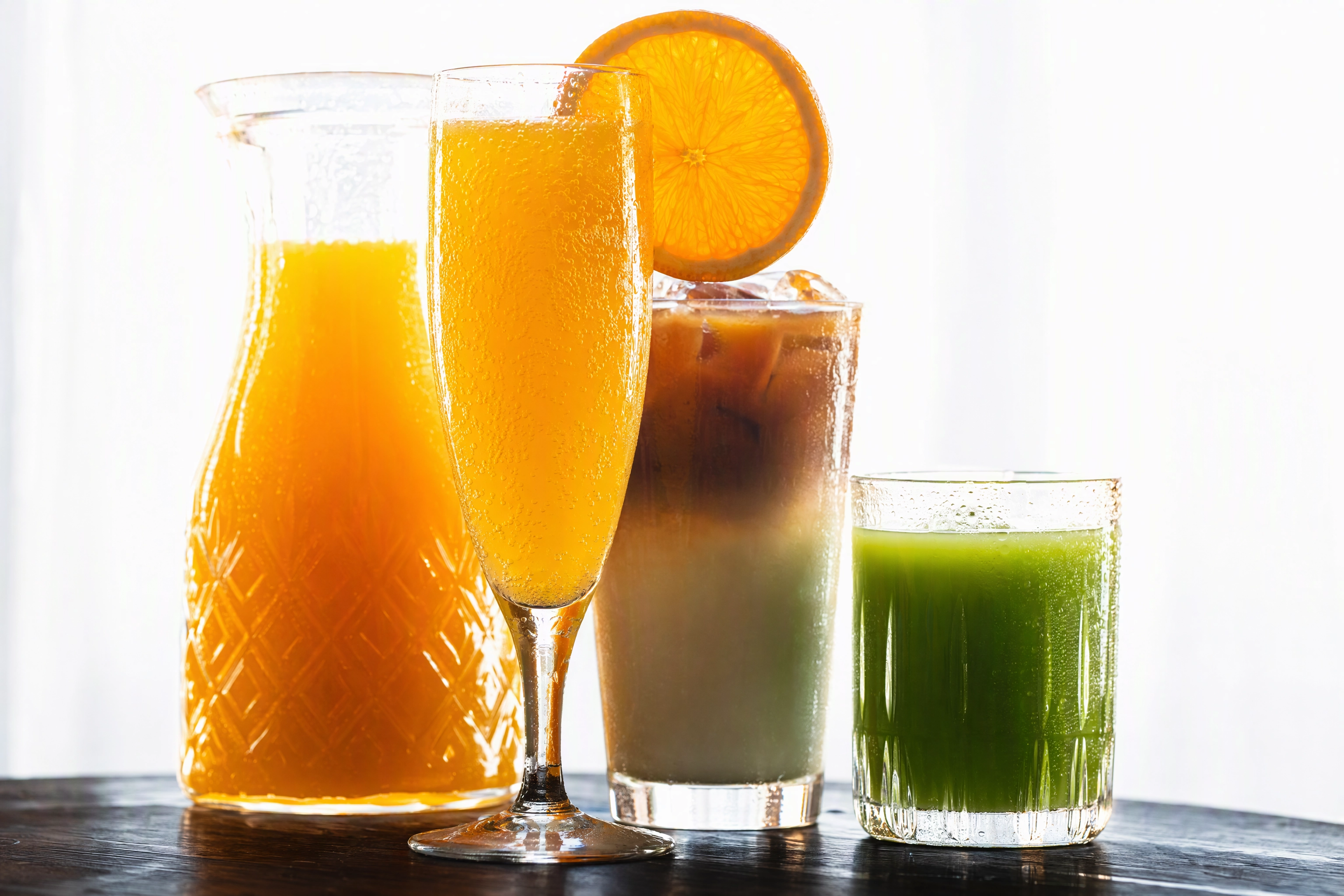 Backlit brunch drinks cluster with mimosa, iced latte and green juice glowing against a bright window