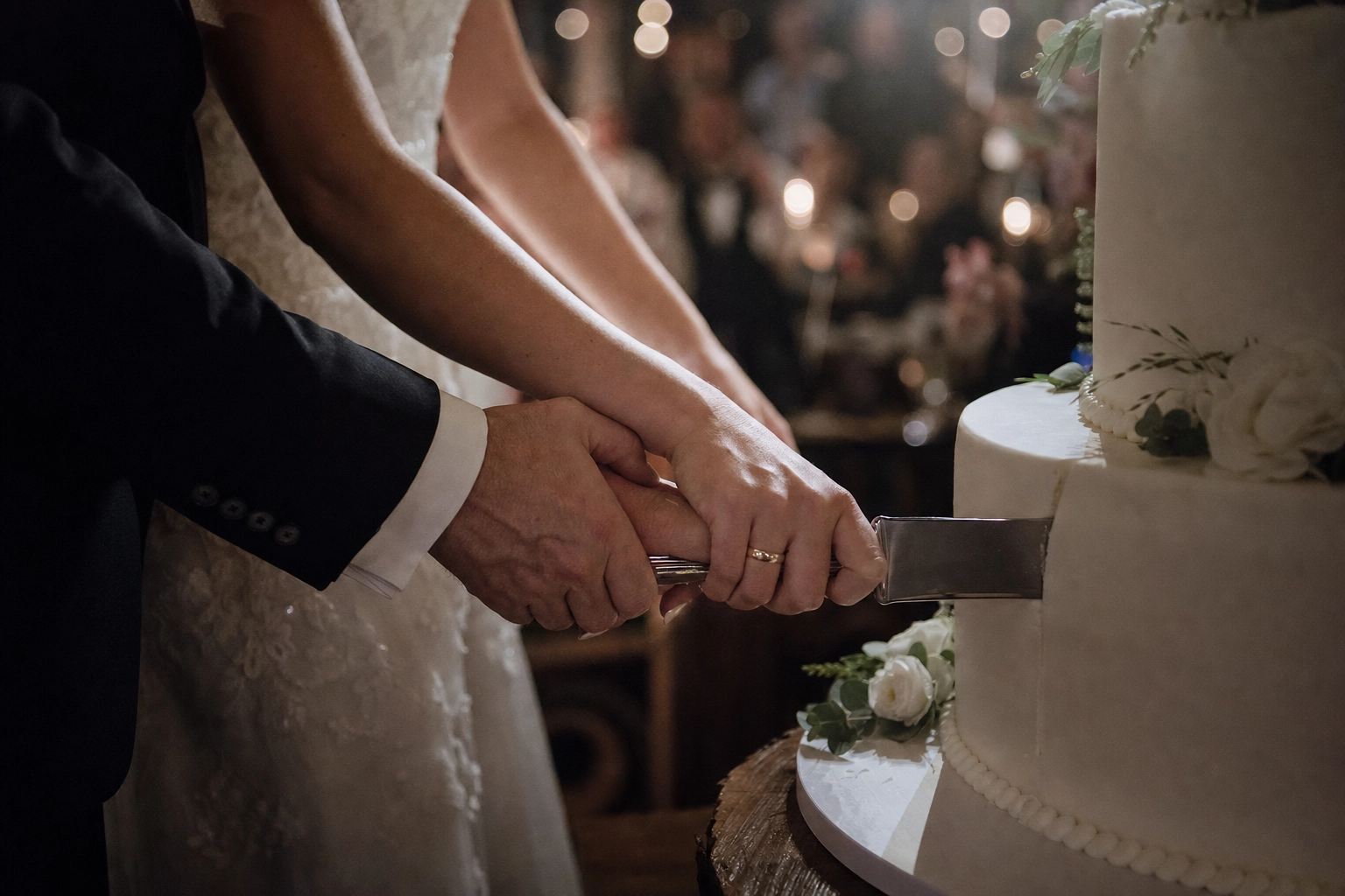 Couple's hands on knife mid-cut into white wedding cake with blurred reception background — cake cutting moment