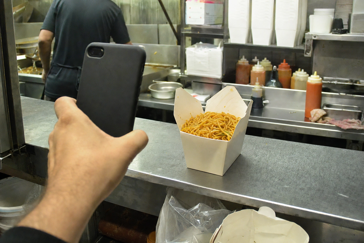Hand holding a smartphone to photograph a dull carton of chow mein on a steel counter under harsh kitchen fluorescent light