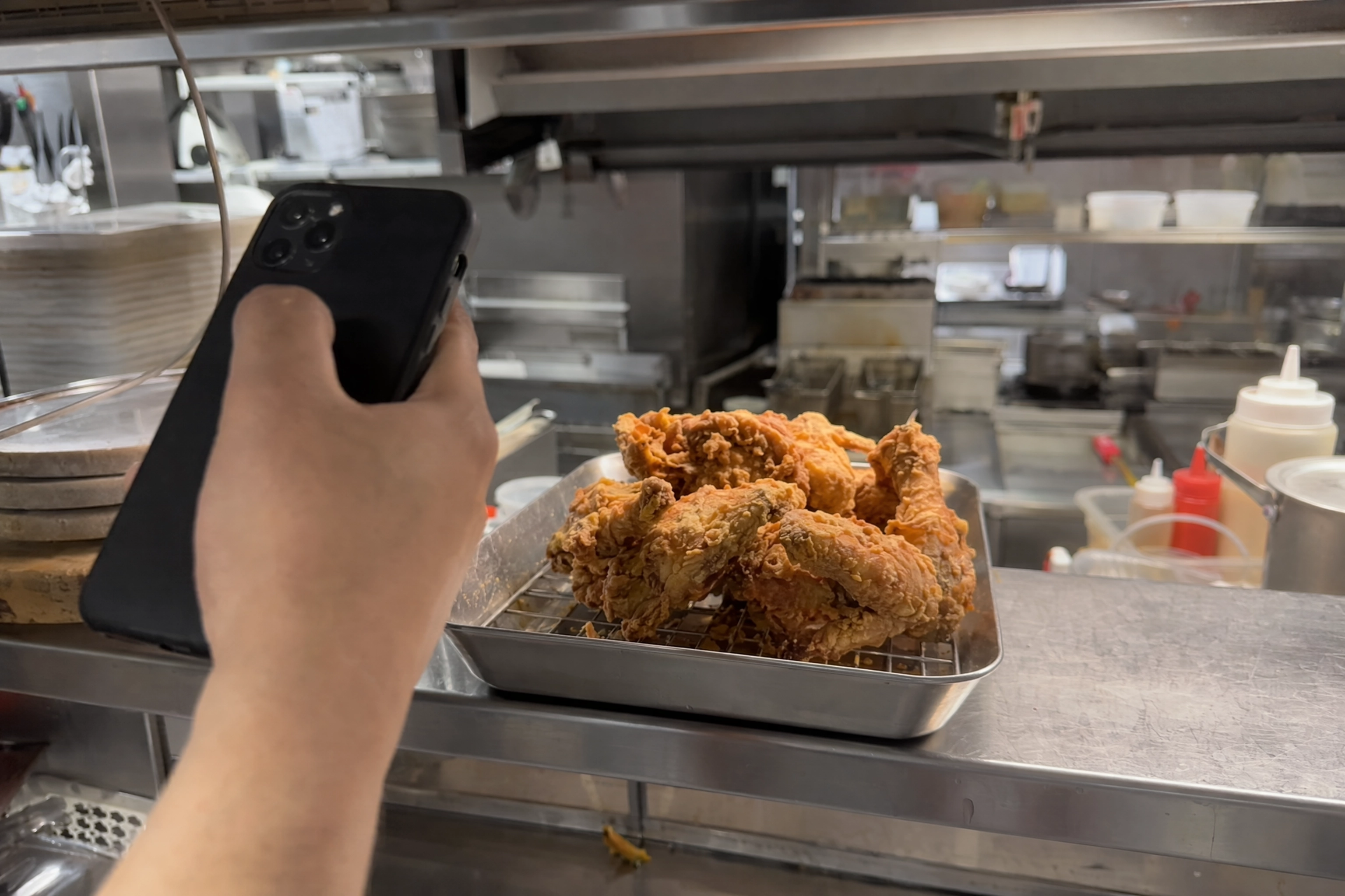 A hand using a smartphone to photograph a tray of fried chicken on a stainless kitchen pass under flat fluorescent light