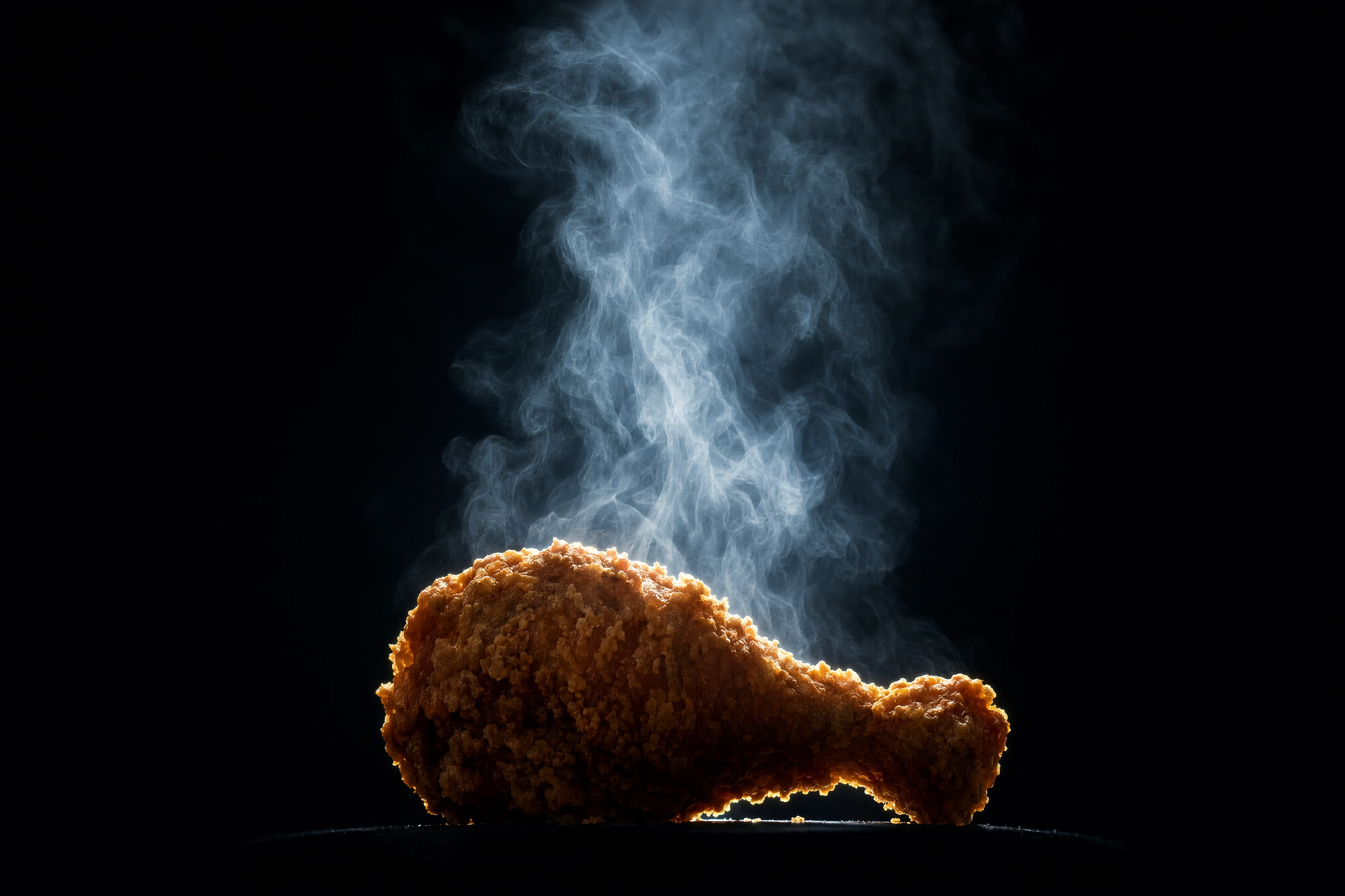 Hot fried chicken drumstick backlit against a black background with a glowing plume of steam rising and separating