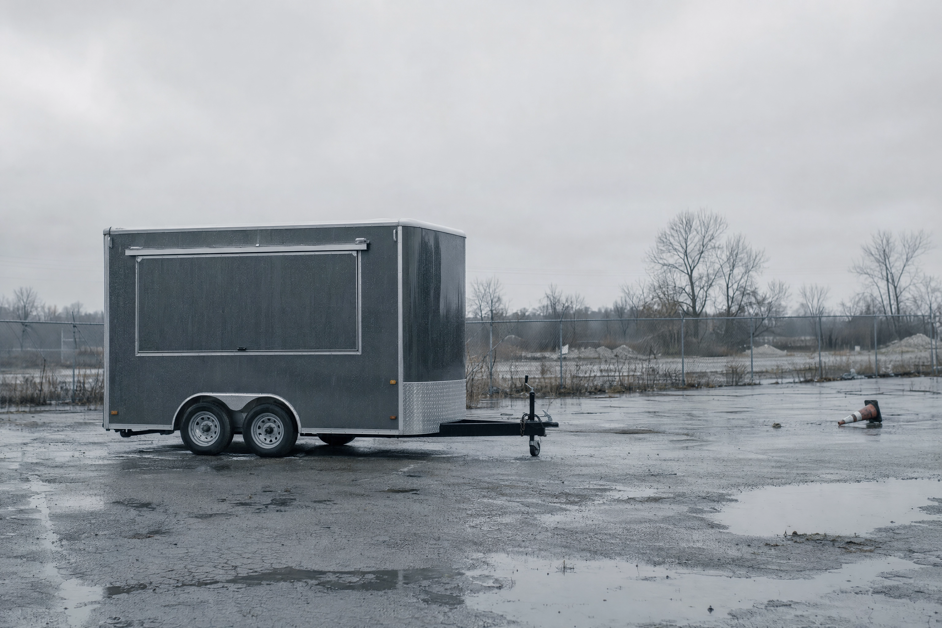 Shuttered food trailer alone on a wet empty lot in flat grey winter light, showing food truck seasonality risk