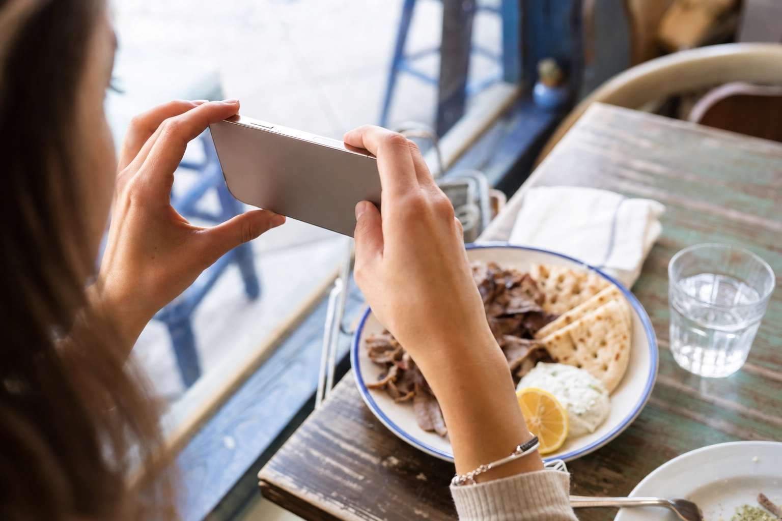 Hands holding a phone to photograph a Greek gyro platter by a window in a real taverna, behind the scenes