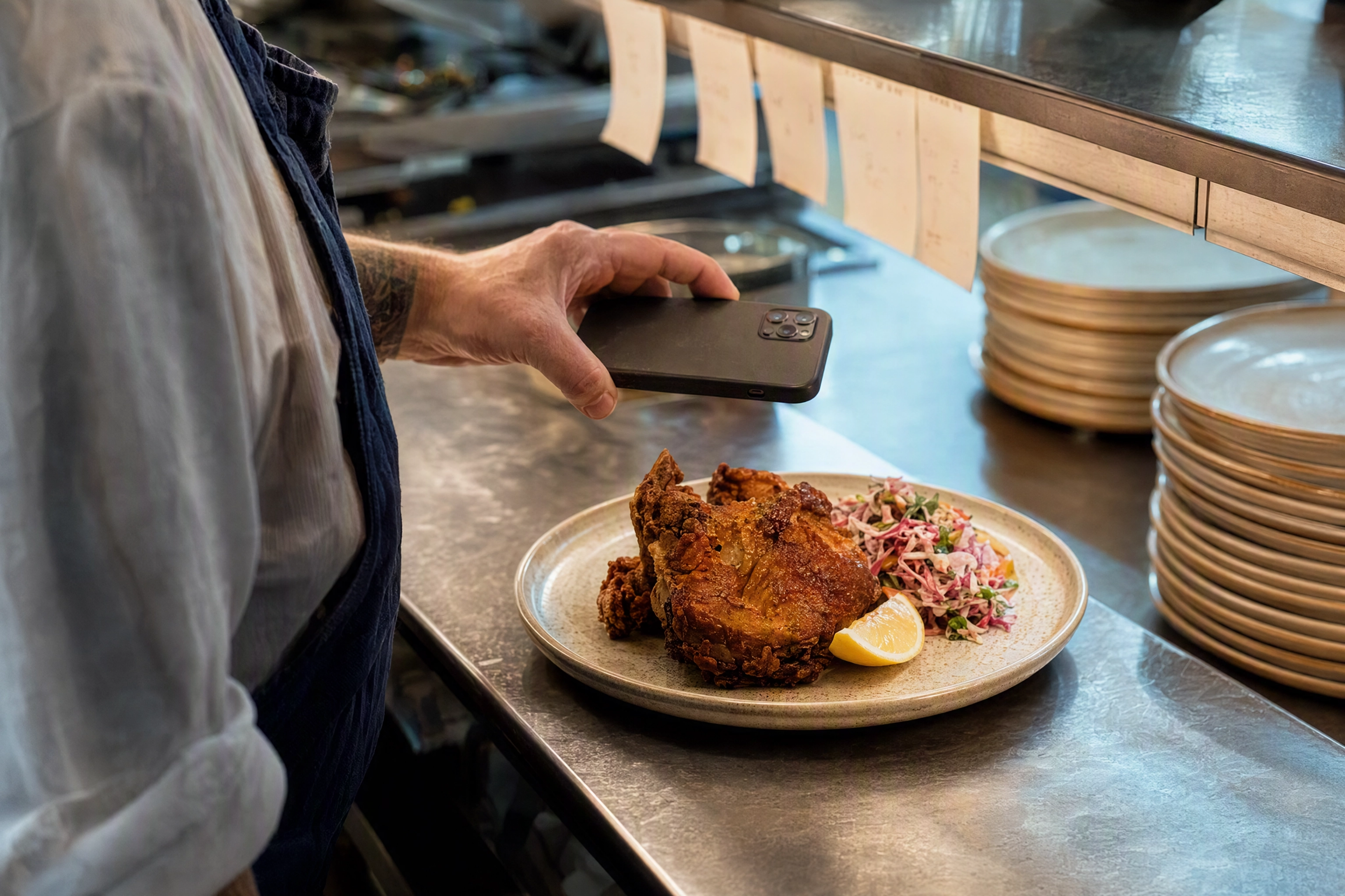 Chef's hands holding a phone back-on above a plated dish at a stainless kitchen pass, capturing a menu photo for the Grubhub merchant portal