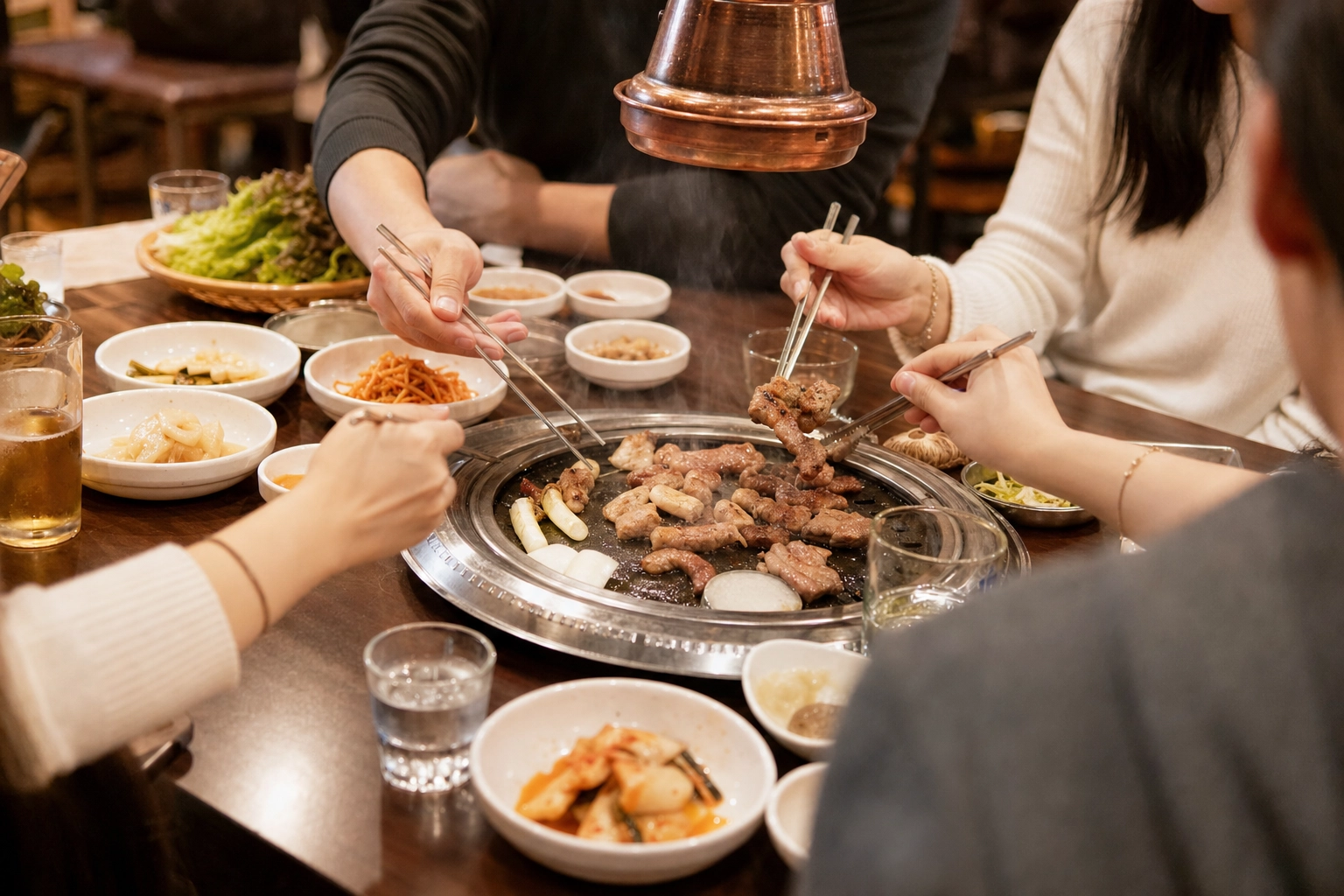 Several diners' hands with metal chopsticks sharing grilled meat and banchan around a sizzling Korean BBQ grill