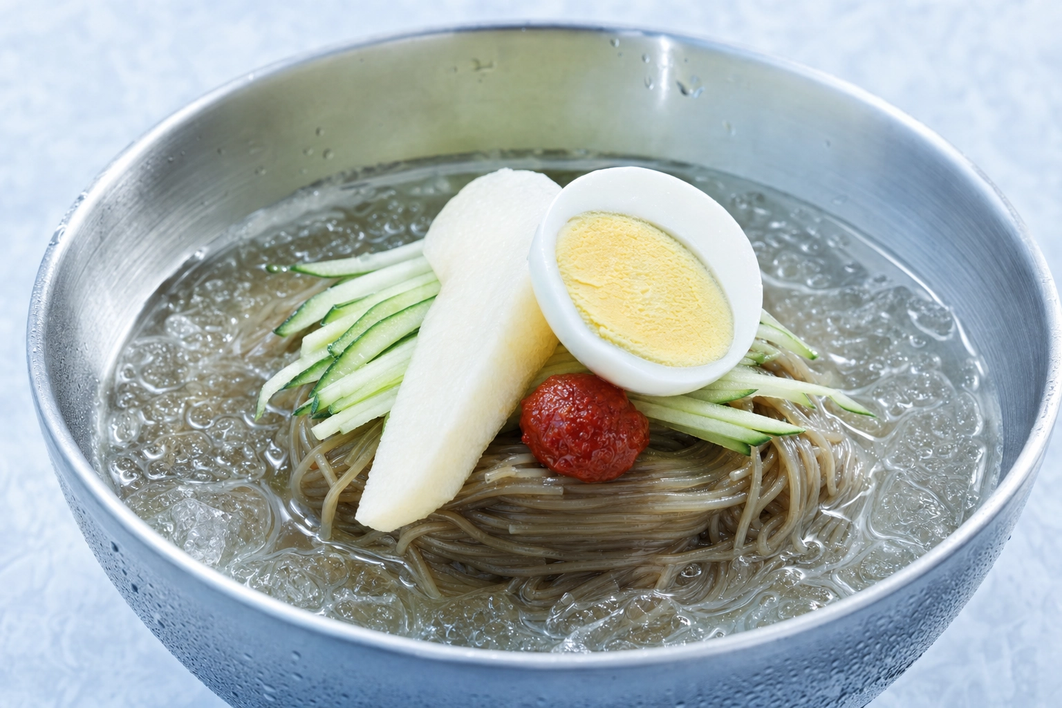 Korean naengmyeon cold buckwheat noodles in icy broth with cucumber, pear and boiled egg in a steel bowl