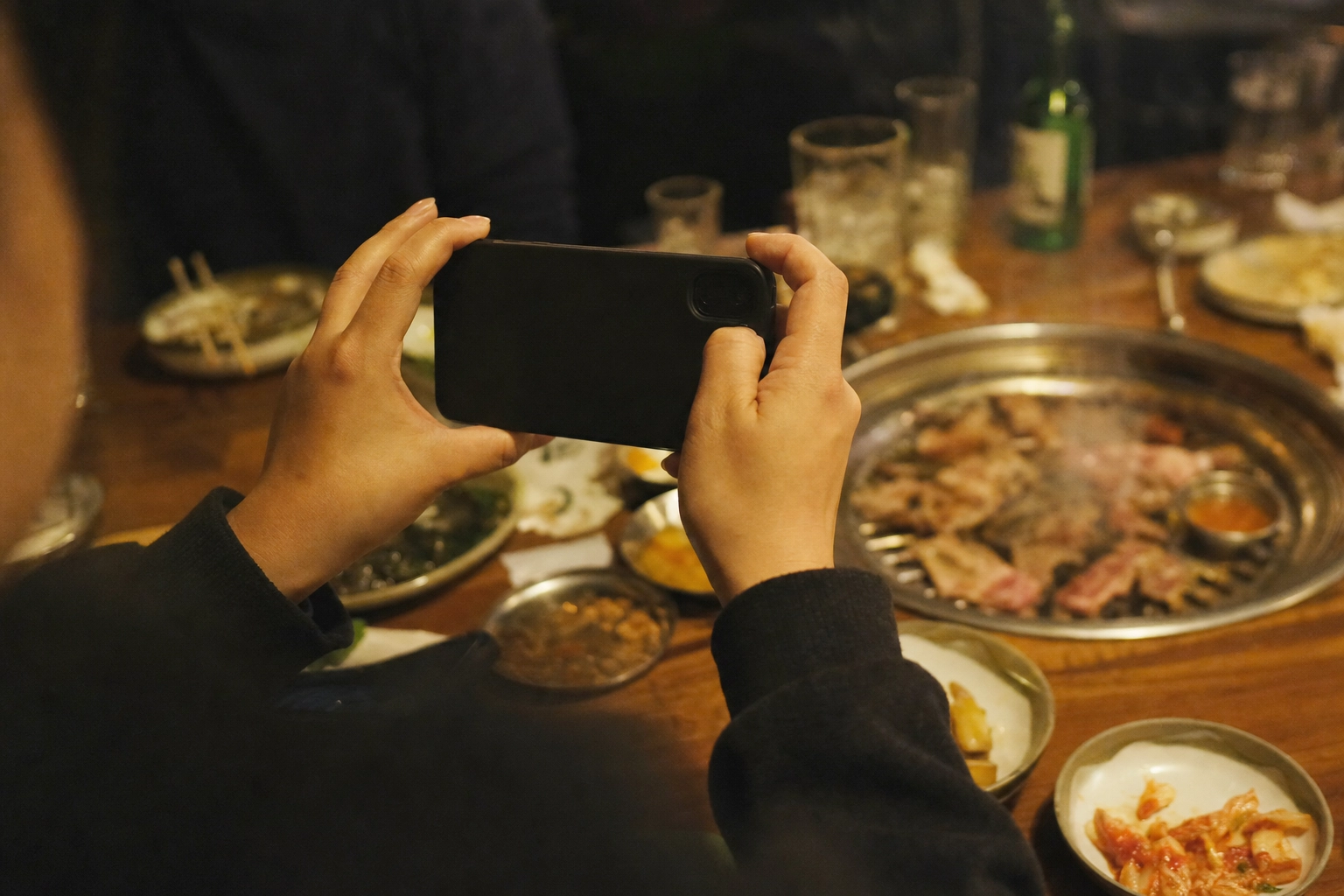 Hands holding a smartphone photographing a sizzling Korean BBQ grill in a dim, warm-lit restaurant booth