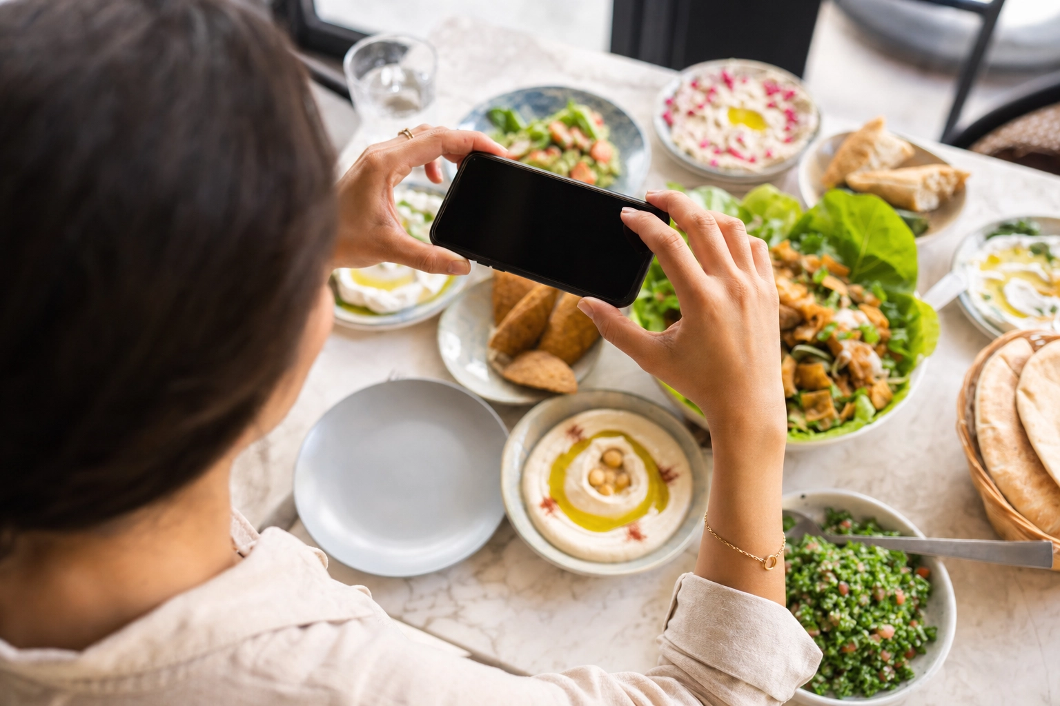 Person holding a smartphone directly above a Lebanese mezze spread to shoot an overhead flat-lay photo