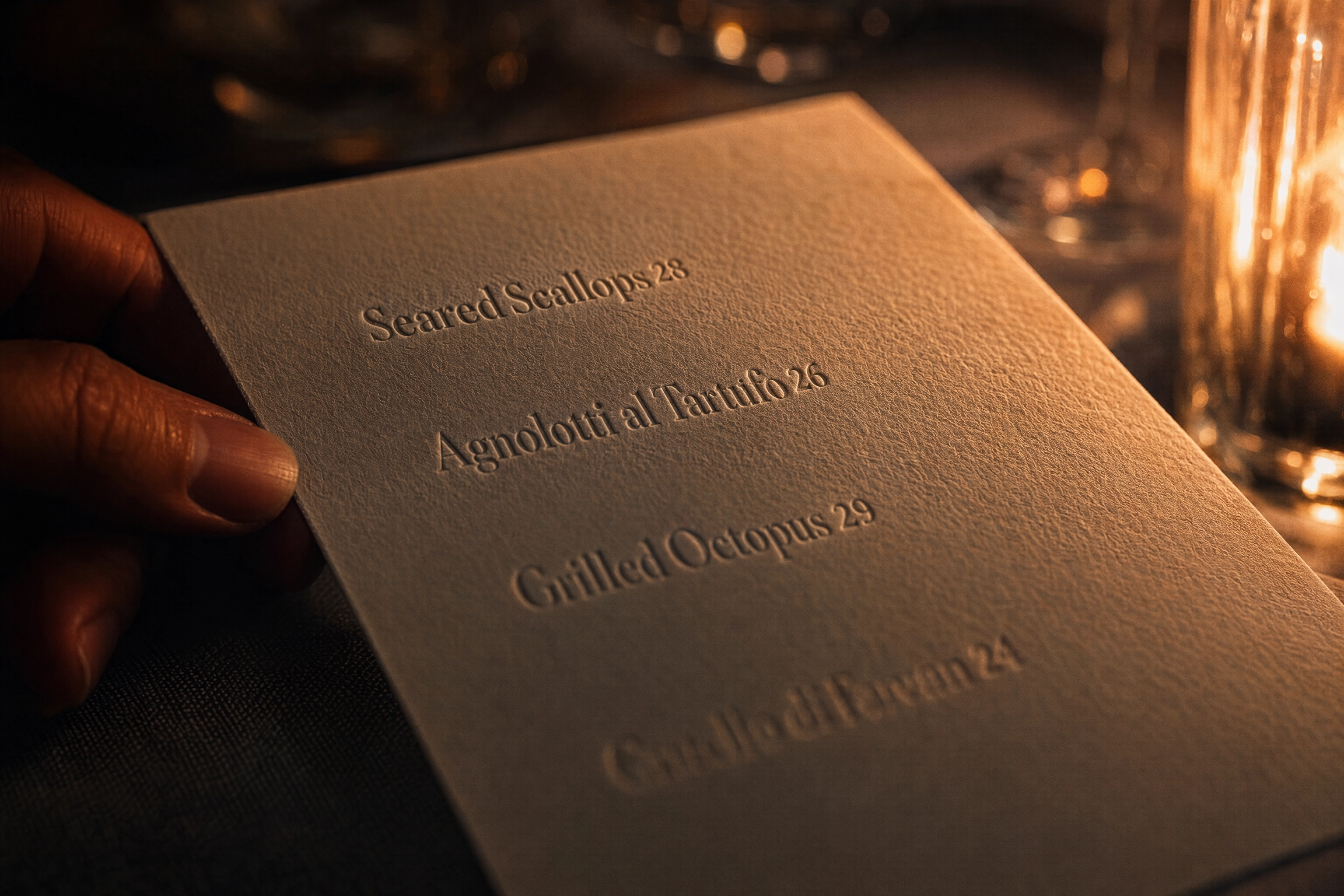Candlelit close-up of an embossed menu card with plain numeral prices and no currency symbols beside a diner's hand