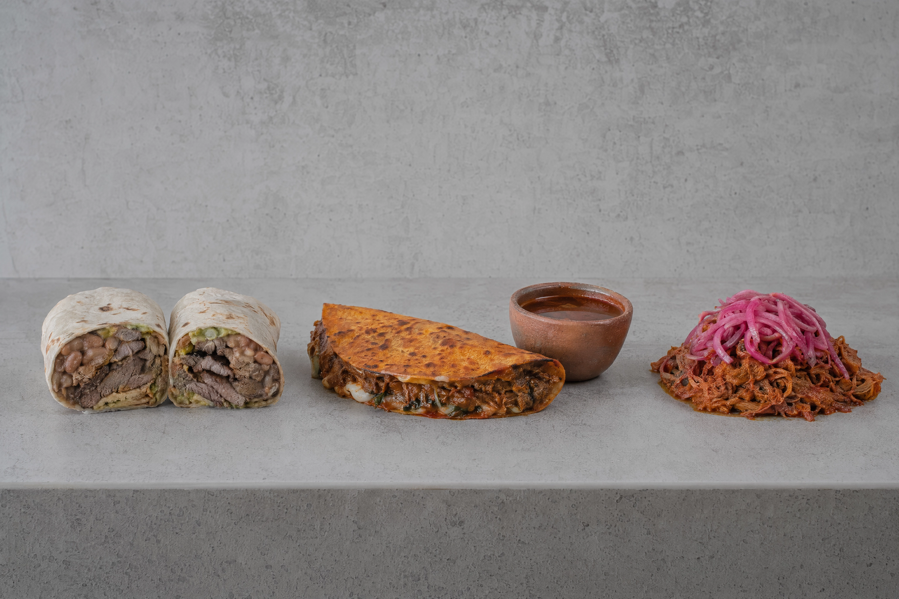 Three regional Mexican dishes compared side by side on concrete: Sonoran flour tortilla burrito, Jalisciense birria with consomme and Yucatecan cochinita pibil