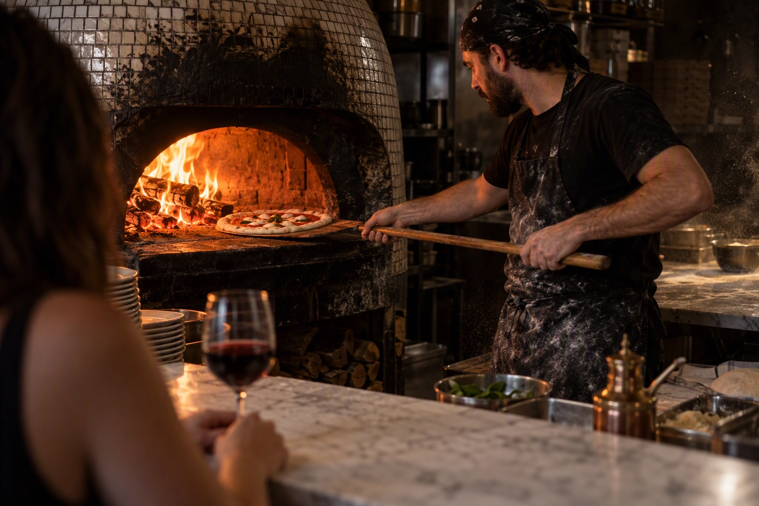 Pizzaiolo sliding a Neapolitan pizza into a wood-fired oven as a dine-in guest watches from the counter, a pizza marketing idea