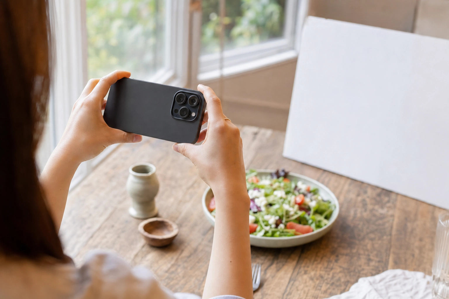 Hands holding a smartphone from behind to photograph a styled salad by a bright window with a white foam-board reflector
