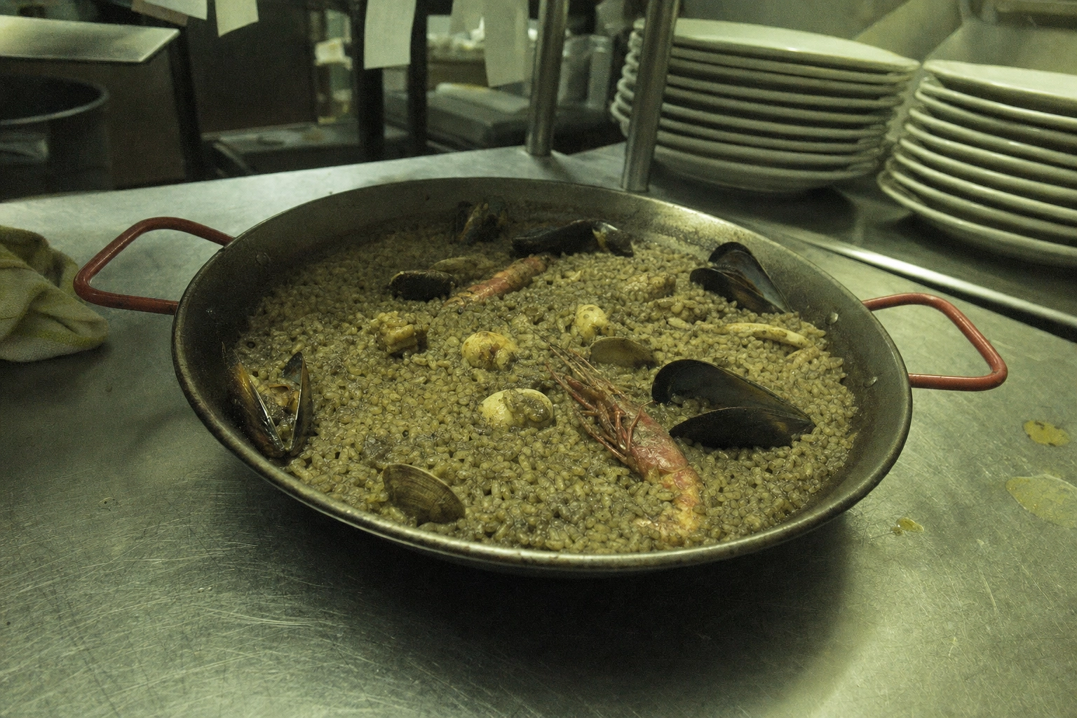 Dull amateur phone photo of paella under flat fluorescent kitchen light on a cluttered stainless pass, the before shot