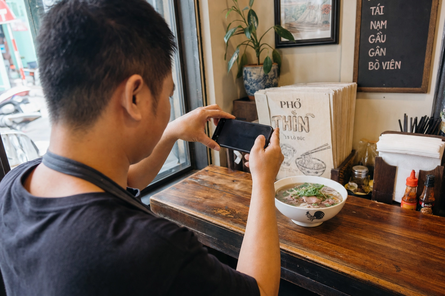 Pho shop cook photographing a fresh bowl of pho with a smartphone on a wooden counter in soft daylight