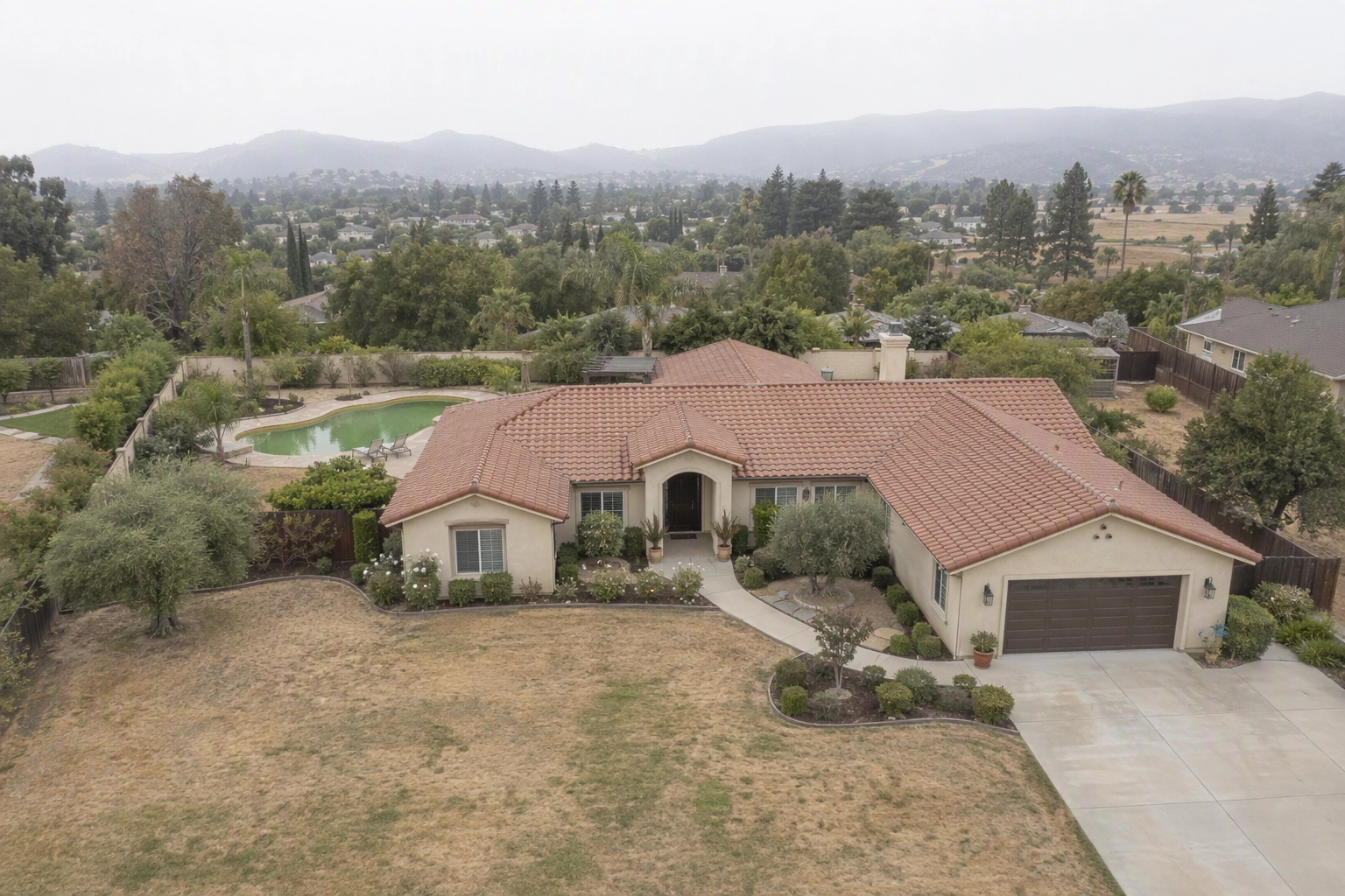 Unedited drone photo of a ranch home under a white overcast sky with a drought-browned lawn and cloudy pool