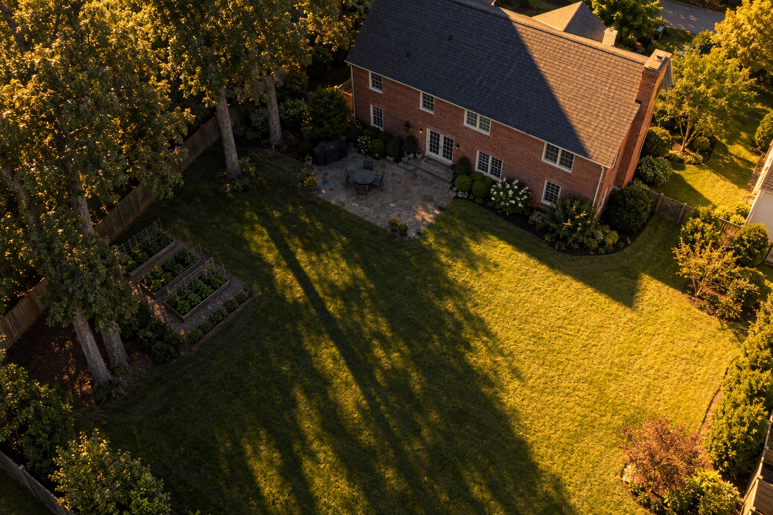 Late-afternoon drone view of a brick Colonial backyard where long oak shadows hide half the lawn and patio