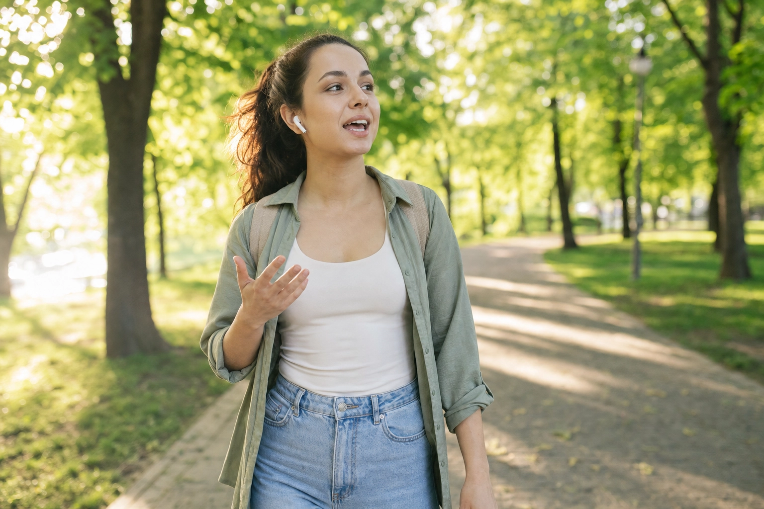 Young woman walking through a sunny park speaking a new language aloud with wireless earbuds during outdoor speaking practice